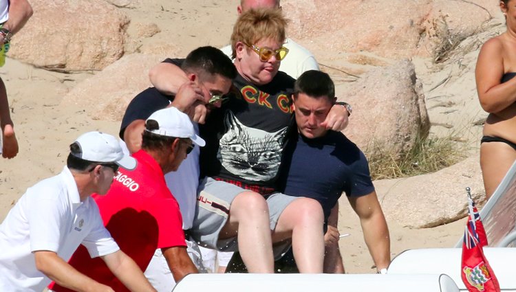 Elton John siendo ayudado para subirse a una pequeña embarcación en Cerdeña