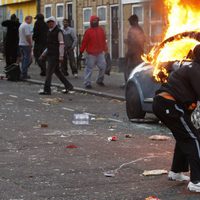 Un joven protagonista de los disturbios de Londres junto a un coche en llamas
